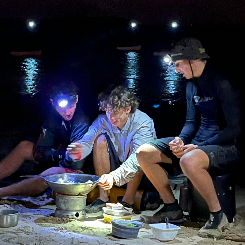 Three men sit on the seashore, enjoying camp cooking during a sea trek at Camp Fairbridge