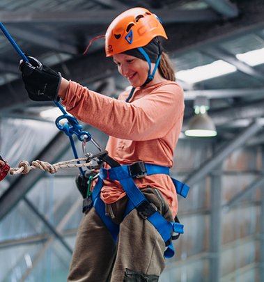 A kid wearing a harness and helmet smiles, enjoying an indoor activity at Camp Fairbridge