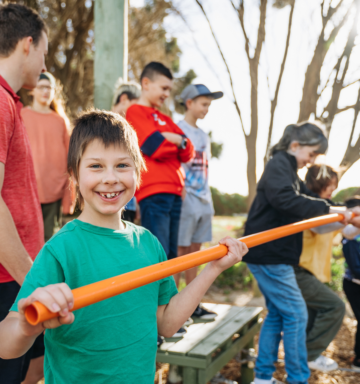 A boy holds an orange pole, surrounded by children, exploring nature in an outdoor classroom setting at Camp Fairbridge