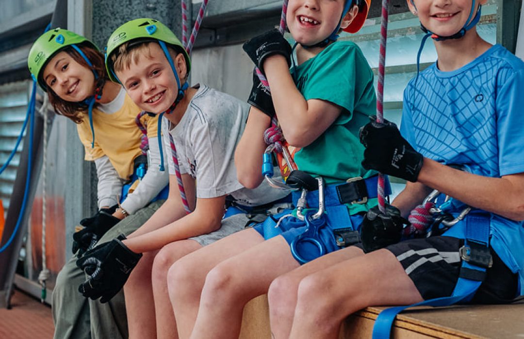 Four children wearing helmets and harnesses hold ropes, ready for an outdoor adventure at Camp Fairbridge Four children wearing helmets and harnesses hold ropes, ready for an outdoor adventure at Camp Fairbridge